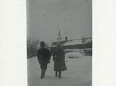 Två barn går i snön med ryggarna åt fotografen mot ett timrat hus. Framför det ligger vedtravar och en eka, bakom huset sticker en kyrkspira upp. (Samma motiv som bild E5436)