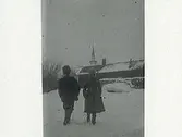 Två barn går i snön med ryggarna åt fotografen mot ett timrat hus. Framför det ligger vedtravar och en eka, bakom huset sticker en kyrkspira upp. (Samma motiv som bild E5436)