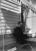 Tonsättaren Ruben Liljefors sitter vid husvägg och läser tidning, sannolikt i Sverige