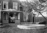 Svens Olle med döttrarna Anna, Britta och Kristina, klädda i folkdräkt, står på veranda till bostadshus, framför huset finns en fontän, Svensgården, Dalarna, midsommar 1919