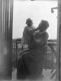 Kvinna sitter på balkong med liten flicka i famnen, sannolikt i Sverige