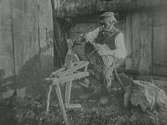 Henrik Bengtsson i södra Björsgård sitter på en 'träskomärr' - en anordning man använde vid träskotillverkning - och tillverkar en korg. Han har en pipa i munnen, träskor och sitter vid en skiftesverkslada med halmtak. (Se även bildnr VMA 6719_2.)