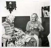 Alma Olsson och Maria Johansson sitter bredvid varandra i en soffa i Marias finrum. Alma har en kofta från Bohus Stickning, Blå Randen - formgivare Anna-Lisa Mannheimer Lunn - och Maria stickar på en kofta till samma företag. Mellan sig har de garner och mönsterdiagram till olika mönster som ingick i Bohus Sticknings kollektioner. Båda stickade åt Bohus Stickning, bilden är tagen inför Marias 80-årsdag och publicerades i Strömstads tidning 16 november 1957.
