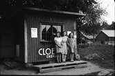 Tre flickor står vid en liten kiosk med Cloetta-reklam. I bakgrunden en gård vid landsvägen.