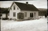 Fageråkras lanthandel Filialen i vinterskrud. Handlare Ottosson står i butiksrock vid entrén, på typiskt butiksmanér förlagd till en avskuren gavel. Till vänster syns en liten lastbrygga och intill den ett räcke att parkera hästen vid.