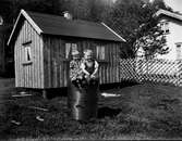 Två små barn, Stina och Olle Olsson i Albäck, sitter på en tunna vid en lekstuga. Mellan den och bostadshuset står ett staket.