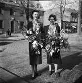 Två studentskor behängda med otaliga blombuketter vid Varbergs kyrka. I bakgrunden syns en mor med barn och barnvagn vid Televerkshuset.