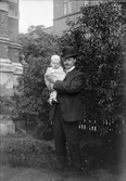 Ung man med litet barn, sannolikt en av sönerna till Ruben och Christiane Liljefors, i famnen ute på gård i stadsmiljö, sannolikt i Sverige
