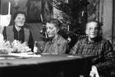 Tre äldre kvinnor sitter vid en julgran i finrummet. Granen är pyntad med stearinlljus, kulgirland och olika julgranskulor. På bordet skymtas en julängel som står bland glitter och en liten tomtefigurin (suddigt i förgrunden).