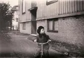 Thore Kristoffersson cyklar på trehjuling utanför huset Frölundagatan 20-22 i Mölndal, ca 1948-1949. Porten till nr 22 syns på bilden likväl som huset 