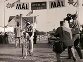 Cykel-SM i Mölndal, år 1956. Cyklisten Per Landén korsar mållinjen vid seniorernas 5-milslopp fredagen den 3 augusti. Starten började klockan 15 på Skedegatan. Målet var på Kungsbackavägen (Brandahls). Banans sträckning: Mölndal-Kungsbacka till vändpunkten söder om Hanhals kyrkoruin och åter till Mölndal. Loppet var Per Landéns sista. Segrade gjorde Herbert Dahlbom före tvåan Gunnar Lindgren och Gunnar Göransson på bronsplats.