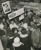 Interiörbild. Svenskt i Topp. Gustavsbergs Fabriker demonstrerar tillvägagångssättet vid porslinsdekor. Turné ute i landet, sannolikt i samarbete med kooperationen i Sundsvall. Plats: Varuhus.
Personer: Okänd dekormålare från Gustavsbergs Fabriker och okända personer..