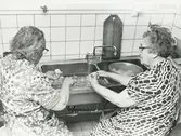 Interör. Gustavsgården i Gustavsberg 1976.Två damer skalar potatis inför den årliga kalaset, en sillsexa, som gårdens gäster själva förbereder.
Personer: Katarina Höst och Elsa Strömberg