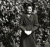 Anna-Lisa i Hugalyckan står framför syrenerna i hatt och kappa över konfirmationsklänningen. Hon håller i psalmboken med vita handskar.