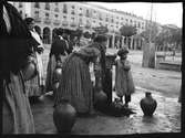 Spanien. Kvinnor och barn vid en vattenpump intill ett torg. Krukor fylls med vatten.