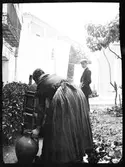 En kvinna som lyfter en vattenkruka betraktas av en man i bakgrunden. Troligen Ronda, Spanien.
