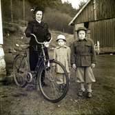 Jenny Lidberg står på gården med cykeln och barnen Elly och Leif. Cykeln har en extra sits bakom styret. De är klädda för en kylig dag med öronlappsmössor, varma jackor och vantar.