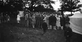 En uppklädd folkskara med många kvinnor har samlats i en ekbacke med uppspända tält. Vad man samlats för att beskåda framgår inte av bilden, men den visar på 1930-talets klädmode med många klockade hattar.