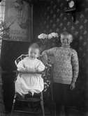 Två barn i ett möblerat rum, troligtvis 1920-30-tal. Det minsta barnet sitter i en barnstol och bredvid står en pojke.