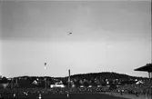 Fotbollsmatch på gamla Ryavallen i Borås, uppförd 1941. Ett par knippen med ballonger stiger till väders, en orkester spelar medan ena laget springer in på plan. Bakom stadion syns villabebyggelsen och en skorsten.