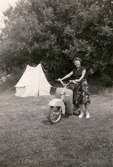 Karin Hasselberg står vid sin scooter, ett tält syns i bakgrunden. Ca 1940-1950-tal.