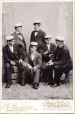 Ateljéporträtt - Sex män iklädda kostym och studentmössa, fyra sitter och två står, några håller rökdon i händerna. Troligen Harald Falk, med handskar i händerna, sittande som andre man från vänster, Uppsala 1899