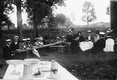 Kvinnor och män, troligen Harald Falk i studentmössa trea från höger med ansiktet mot kameran, sitter vid långbord, bord med skålar och tillbringare i förgrunden. Vid Grindstugan, Uppsala, omkring 1900