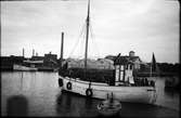 En fiskebåt fullastad med lottor och en eka på släp lämnar Varbergs hamn. Vid kaj ligger att magasin med eternitfasad krönt av en skylt för Borås Wäfveriaktiebolag.