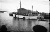 En fiskebåt fullastad med lottor och en eka på släp lämnar Varbergs hamn. Vid kaj ligger ett magasin med välvt tak med lastpallar intill och i bakgrunden syns en Essocistern.