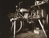 Stilleben inne i fotografen Kloar-Eriks egen stuga där han mekar med en lättviktare. I förgrunden står en lång pall med skruvstäd och olika verktyg uppradade. I bakgrunden syns 98-kubikaren.