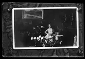 Reprofotografi av ett foto föreställande en kvinna som sitter i ett finrum omgiven av blommor. Troligen i samband med en jämn födelsedag. I fotografens anteckningar står det 