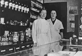 2 kvinnor, Kerstin Landgren och Elsa Landgren, i vita rockar står bakom en butiksdisk och tittar in i kameran. Bakom sig har dom en hylla fylld med glasburkar.
Landgrens Kemigrafiska Affär. Kungsgatan 41.