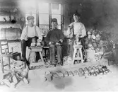 Toffelverkstaden Prästgatan 23, Varberg år 1908. Männen sitter vid sina redskap med spån på golvet och träämnena i en trave till höger. Framför sargen står färdiga tofflor. Från vänster: ägaren till verkstaden, Alfred Johansson, Varberg, Birger Bengtsson och Carl Bengtsson, båda från Varberg.