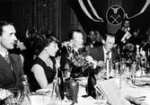 Gäster kring ett restaurangbord vid 10-års jubileet 1954 av Svenska sadelmakare- och tapetserareförbundet avdelning 63 på Stadshotellet i Alingsås.
En servitris med handen vid hakan redo att servera.

Enligt text på bildens baksida är personerna på bilden:
Åke Engqvist
Fru Norell
Karl Erik Norell
Förbundsordförande Gustav Eriksson
Arne Eriksson