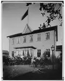 Gruppbild av 34, sittande och stående, kvinnor, män och pojkar framför ett hus. Huset har ett tympanon med texten N, G, T 1888 samt en symbol av ett hjärta korsat med kors och ett ankare. Till höger på bilden ses två cyklar, musikinstrument på gräset och ett småbarn. Stående vid cykel t.h. Fridolf Thorssin. 

NTO:s lokal i kv Hammaren vid Göteborgsvägen.