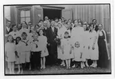 Ett fyrtiotal bröllopsgäster inklusive brudparet i en gruppbild. De står på gården utanför ett hus. Originalet i vykortsformat, bruntonat.
