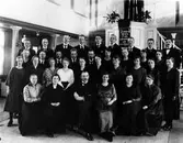 Gruppbild på 34 kvinnor och män i missionskyrkans kyrksal, stående och sittande, framför kyrkans predikstol. Under antependiet en tavla med ett bibelcitat.