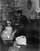 En jultomte står framför en julgran och håller i en dockvagn med en docka i, ett barn står jämsides och blundar.