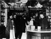 3 män står bakom disken i en speceriaffär och hjälper 2 par kunder. Interiör av Alfred Hedéns affär vid Stora torget.