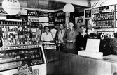 Inifrån Sandbergs speceriaffär. Bakom disken står 3 män, 1 kvinna och 1 tonårspojke. Till vänster en stor glasdisk fylld med olika sillinläggningar.
Kvarteret Storken.