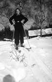 En ung kvinna på skidor med en stuga skymtande bakom backkrönet. Hon är vinterklädd med byxor, raggsockar, pjäxor och ett resårskärp om tröjan.