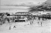 Skidåkare, baracker, järnväg i bakgrunden.