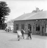 Personal går med postsäckar. Järnvägen kom till Sankt Olof 1901 och drivs fortfarande, dock som museijärnväg. I bakgrunden på bilden ligger ett hotell.