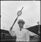 En tågklarerare ansvarar för att tidtabellen och tågordningen hålls och är den som bedömer vilka åtgärder som bör göras vid avvikelser i tågtrafiken. På bilden tågklarerare ger ett signal att tåget får avgå från stationen. Tågklarerare har sommar uniform på sig.