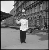 En tågklarerare ansvarar för att tidtabellen och tågordningen hålls och är den som bedömer vilka åtgärder som bör göras vid avvikelser i tågtrafiken. På bilden tågklarerare ger ett signal att tåget får avgå från stationen.  Tågklarerare har sommar uniform på sig.