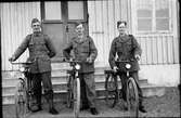 Tre beredskapsmän under andra världskriget står med varsin cykel framför ingången till ett hus, en bygdegård enligt uppgift.