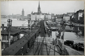 Tåg vid infarten till Mälarstrand. Ställverkshus till vänster i bild. Riddarholmskyrkan och Stadshuset i bakgrunden.