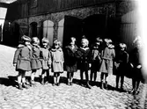 Gruppbild på ett antal barn vid en privat Kindergarten på Kungsgatan, kvarteret Pärlan 10. Brita Hjelmqvist drev kindergarten på övervåningen. 
Från vänster: Kate, Vera, Ingrid, Dockan, Brandt, Britta, Greta, Eva-Stina, Ulla Klinge, Harriet.
