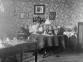 Fröken Maria Elmgren sitter i en soffa och tittar i sin handväska. Rummet har en stormönstrad, blommig tapet. Interiör, kv Magistern, Prästgatan.
