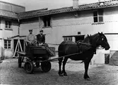 Gårdsplan med träbyggnad i två våningar. På gårdsplanen står en häst med vagn och två män, till vänster åkardrängen Karl Eriksson och till höger Ivar Jonsson.
Gården, i kv Jägaren 11, tillhör Karl-Adrian Svenssons koloniallager och Alingsås Express-åkeri, som ägdes av Per Andersson (Långared). Bakom vagnen syns Alingsås Express-åkeris stallar och kontor med plats för 7-8 hästar. Som mest hade åkeriet ca 20 hästar, då utplacerade på Gunnar Johanssons och Hedens gårdar
